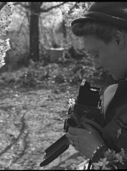 Photographie en noir et blanc de Germaine Kanova, en uniforme de correspondante de guerre, tenant un appareil photo au milieu des fleurs.